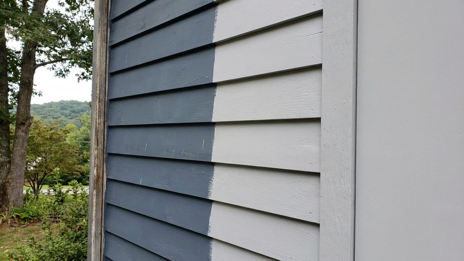 Close-up of fiber cement lap siding on a house wall, half in a deep factory-finished slate blue and half in a lighter freshly painted gray