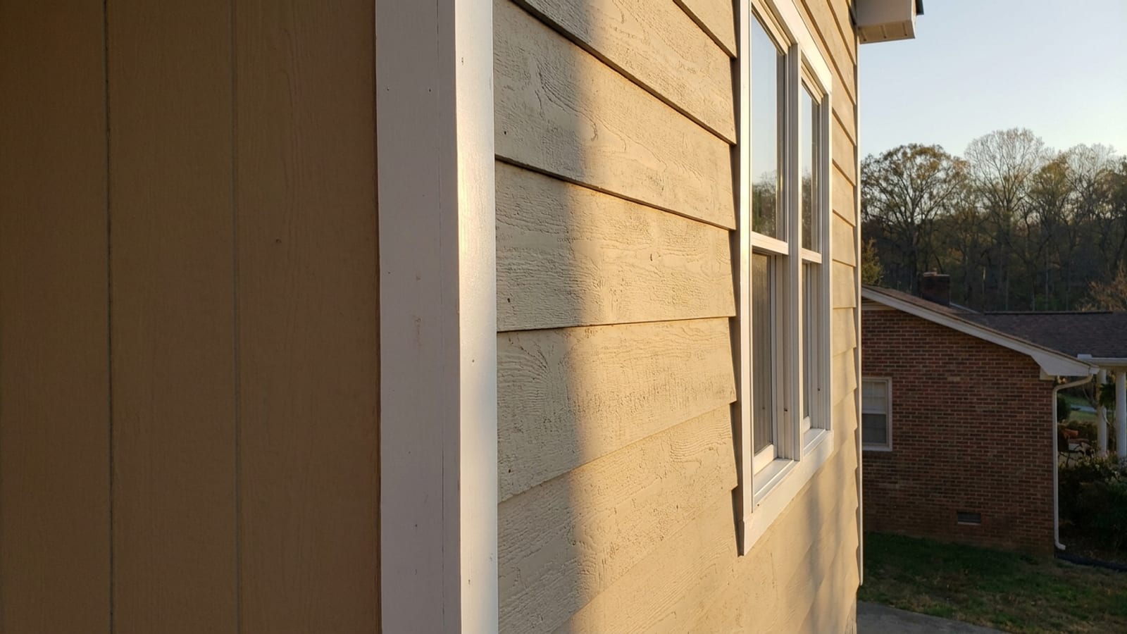 Close-up of a house corner where new lap siding meets a white corner post and window trim in raking sunlight