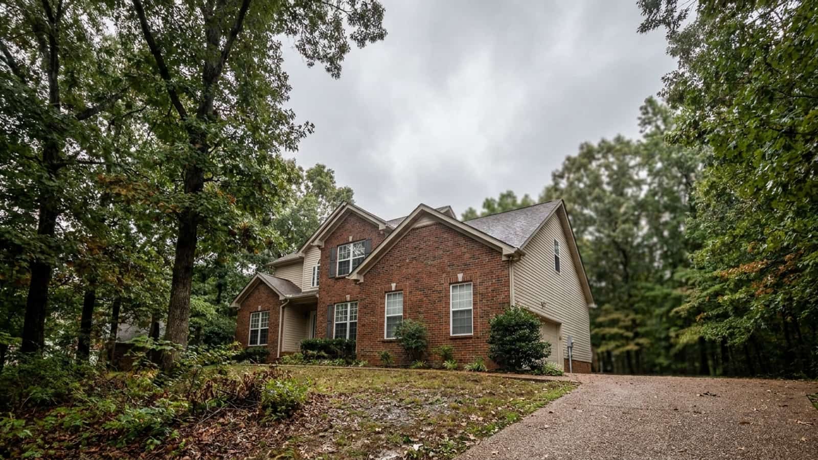 Two-story brick and vinyl suburban home with white double-hung windows under mature oak trees on an overcast day